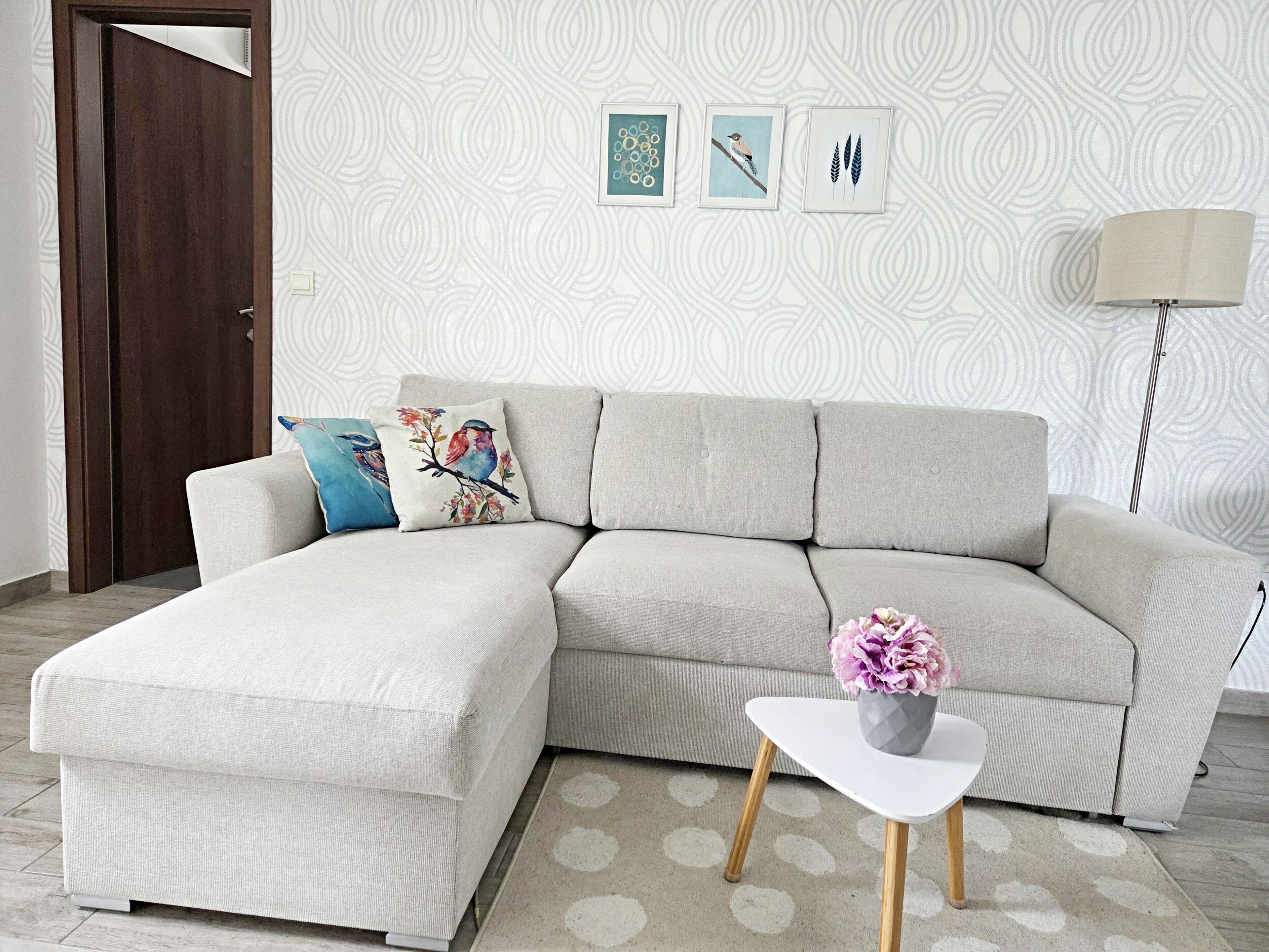 Un salon avec un canapé d’angle, une petite table blanche ornée d’un vase et un mur à motifs.