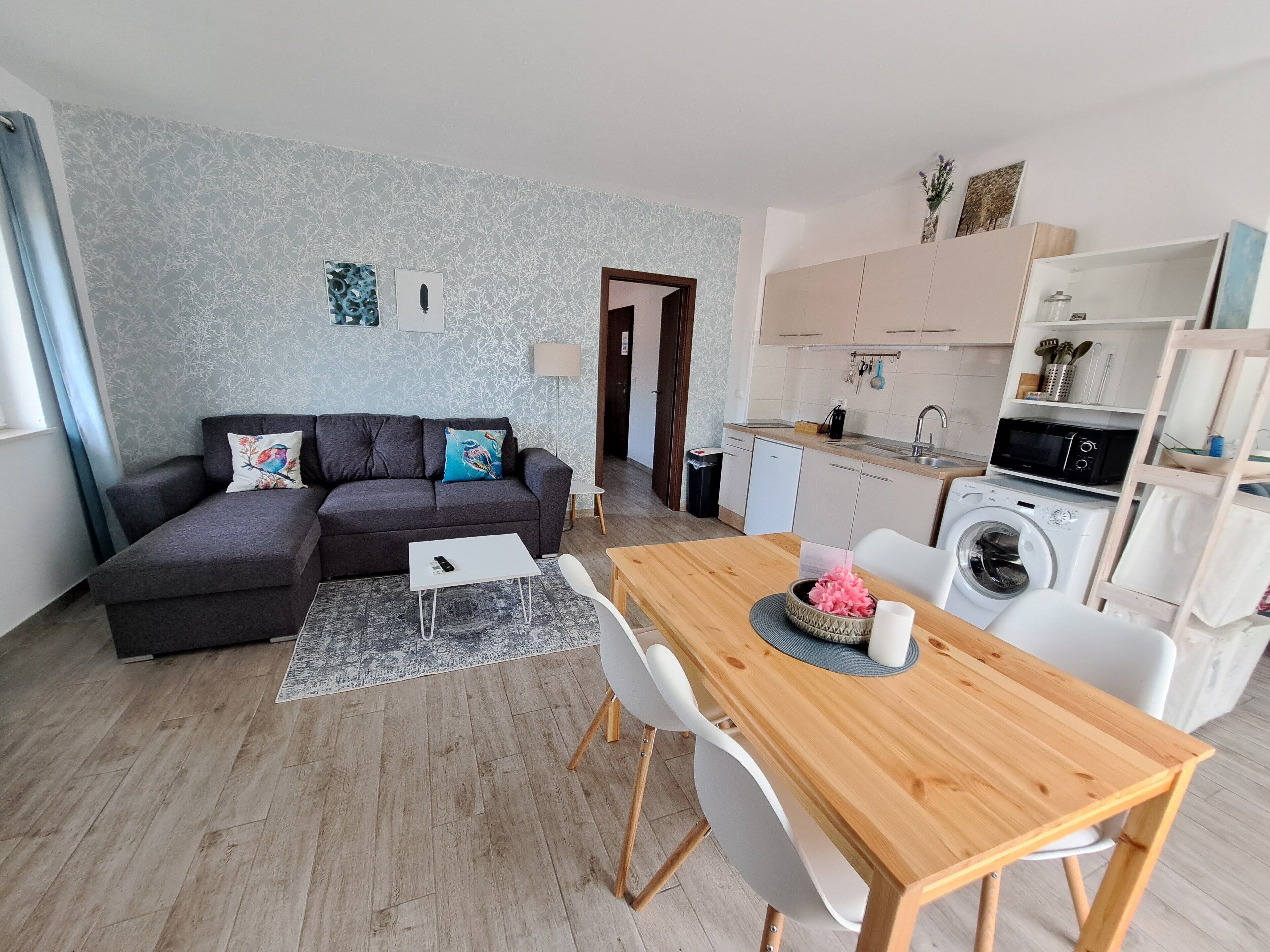 Ein modernes Wohnzimmer mit grauem Sofa, einem Esstisch mit Stühlen, einer Waschmaschine und einer Küchenzeile mit Schränken und einer Mikrowelle.