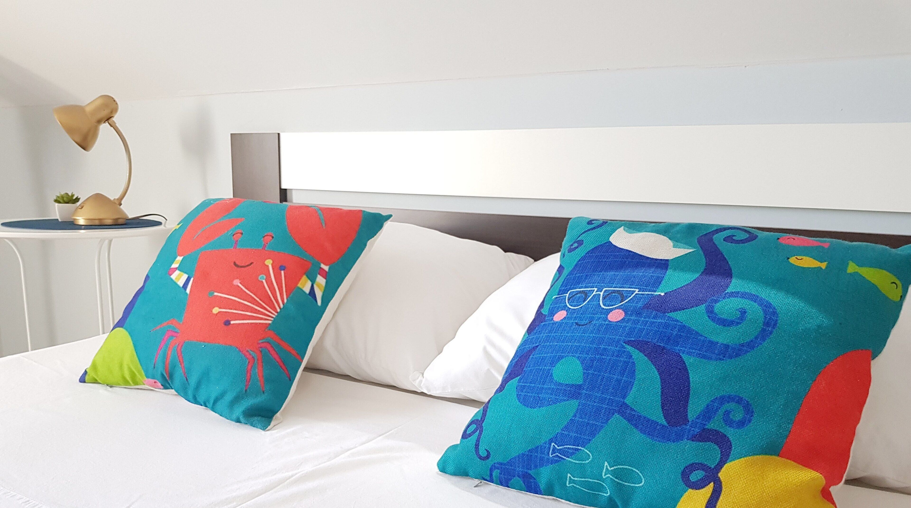Un lit avec deux oreillers colorés, l’un orné d’un motif de crabe et l’autre d’un motif de poulpe, et une petite table avec une lampe et une plante en arrière-plan.