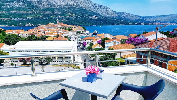 A balcony with two chairs and a table, overlooking a coastal town with buildings and a mountain in the background. - Adriatic Pearl Korcula (Korcula)