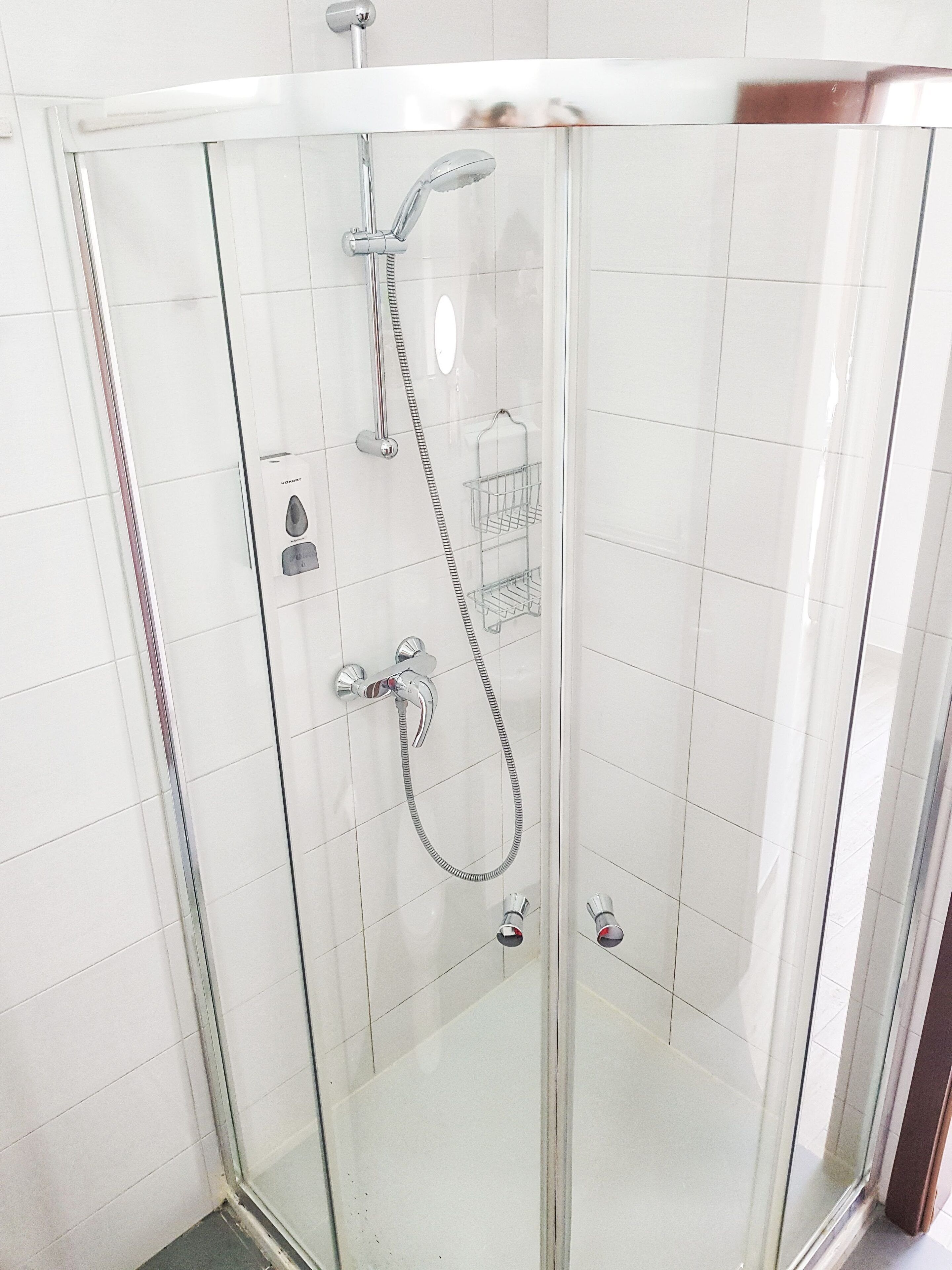 A glass-enclosed shower with a handheld showerhead and a fixed showerhead, a control panel, and a small shelf.