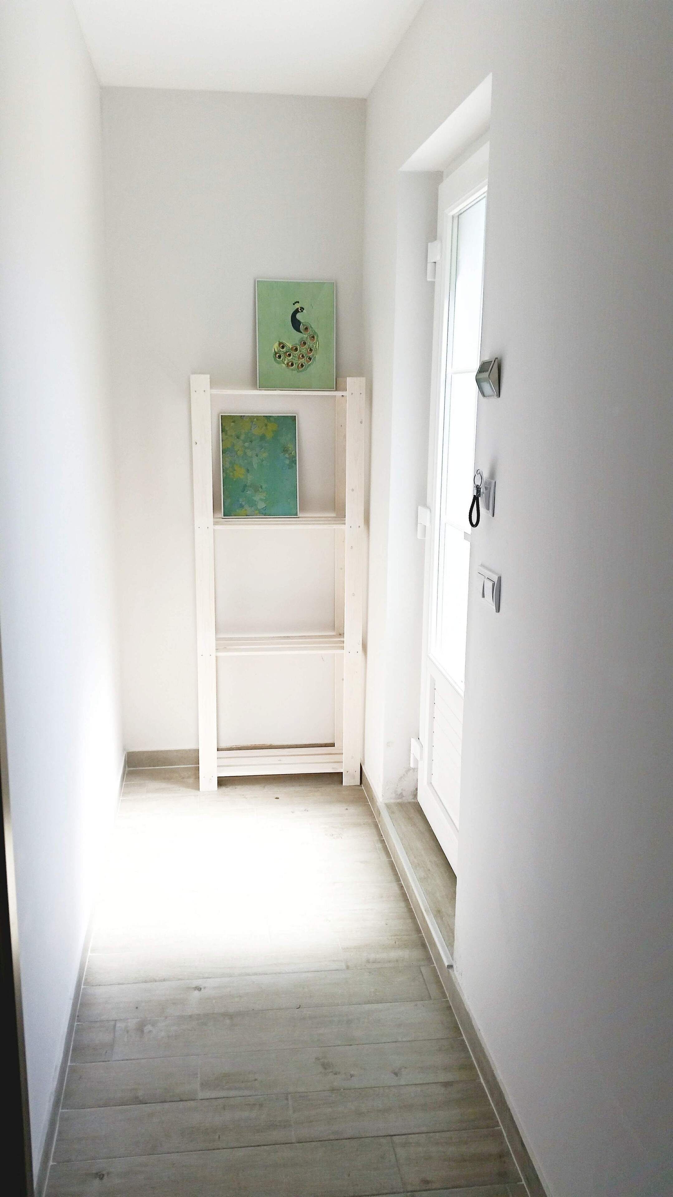A narrow hallway with a white door on the right, a white shelf unit against the wall, and a painting above it.