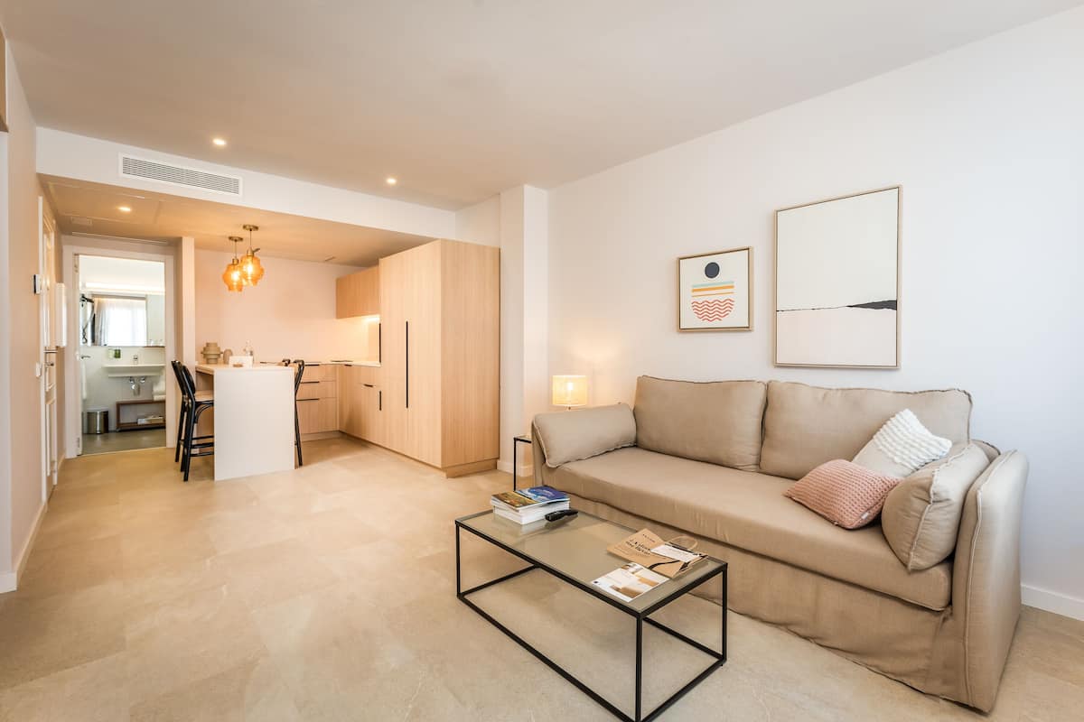 Un salon moderne avec une table basse en verre, un canapé beige et une cuisine en arrière-plan.