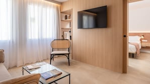 A modern hotel room with a flat-screen TV mounted on the wall, a comfortable sofa, a glass coffee table with books, a wicker chair, and a wooden bookshelf. - ARTIEM Urban Apartments (Mahón)
