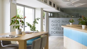 A hotel reception area with a wooden desk, chairs, potted plants, and a large window. - ARTIEM Urban Apartments (Mahón)