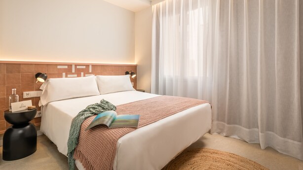 A neatly made bed with a white comforter and pillows, a bedside table with a lamp, and a large window with sheer curtains.