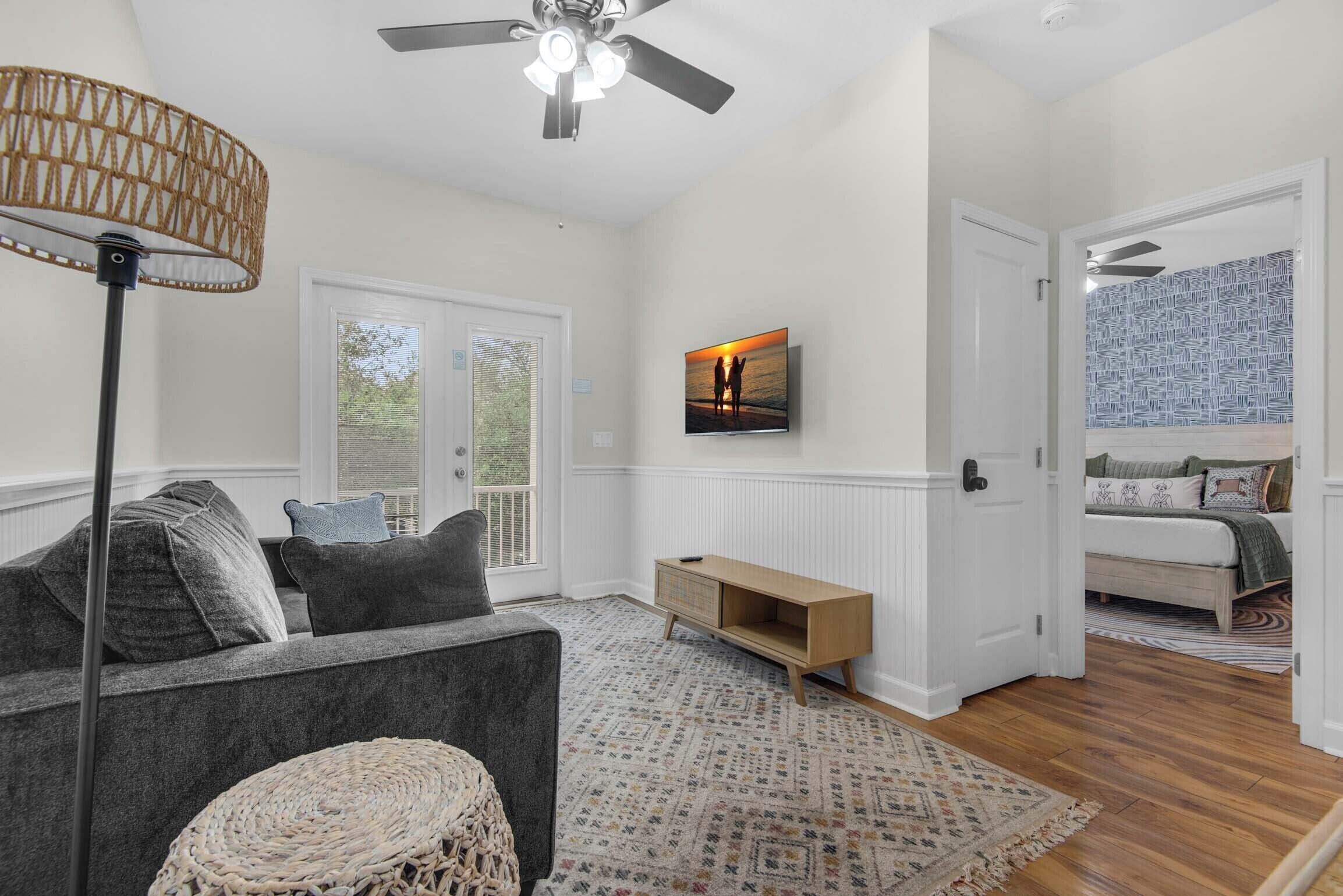 A spacious living room with a gray sofa, a woven ottoman, a wooden console table, and a large TV mounted on the wall.