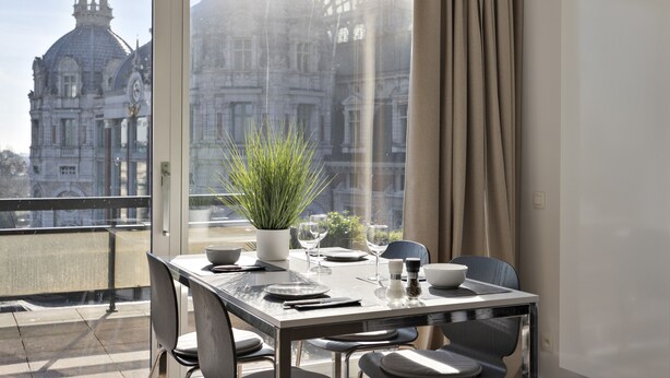 A dining area with a table set for four, a potted plant, and a view of a historic building through a large window. - Luxurious Penthouse, rooftop terrace, spectacular view! (Antwerpen)