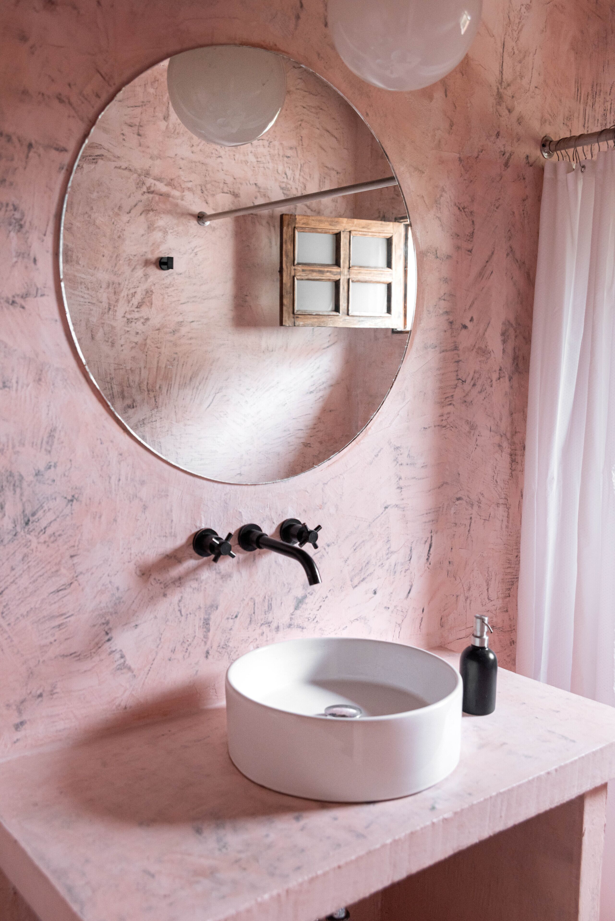 Une salle de bain avec un miroir rond, un lavabo blanc et un distributeur de savon noir.