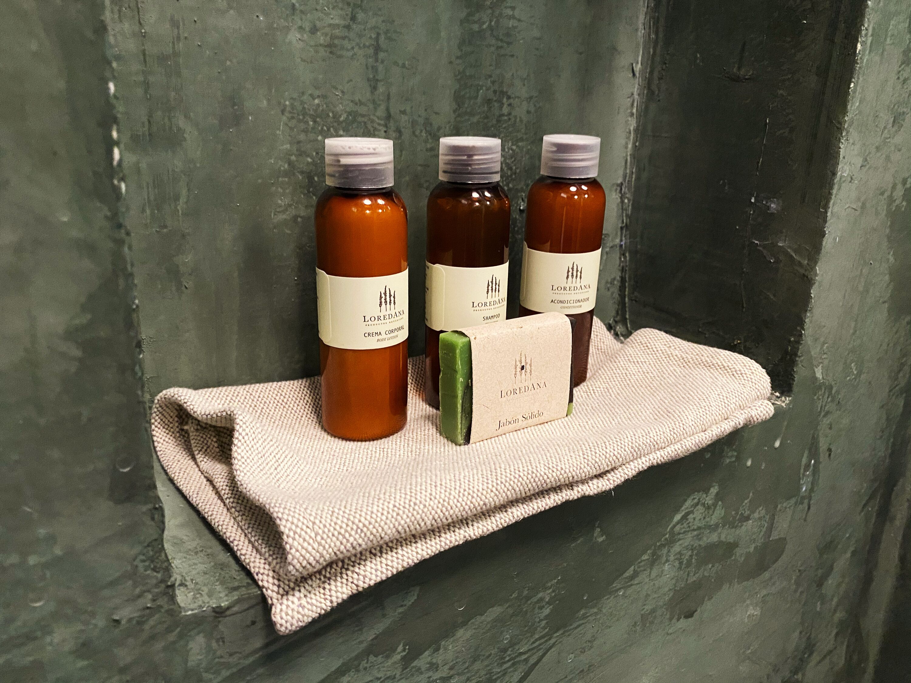 Tres botellas de champú y acondicionador Loredana sobre un estante frente a una pared con textura.