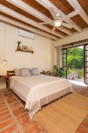 A bedroom with a large bed, a ceiling fan, an air conditioning unit, a wooden headboard, a bedside table, a nightstand, and a view of the outdoors through a glass door. - Casa Ritual (Puerto Vallarta)