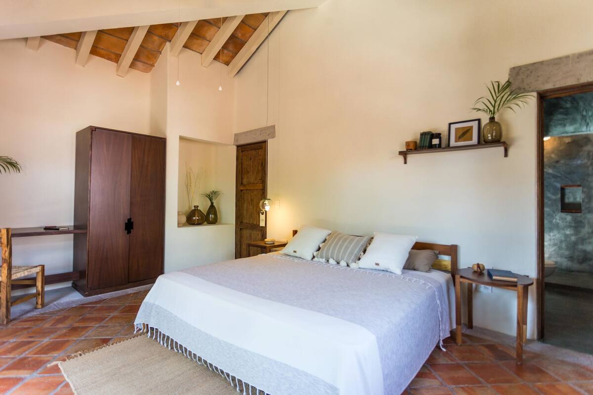 A bedroom with a large bed, a wooden wardrobe, a desk, and a shelf with decorative items.