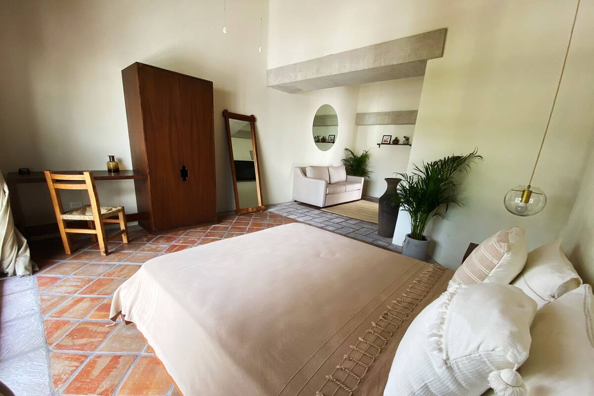 A bedroom with a large bed, a wooden wardrobe, a desk with a chair, a sofa, and a potted plant.