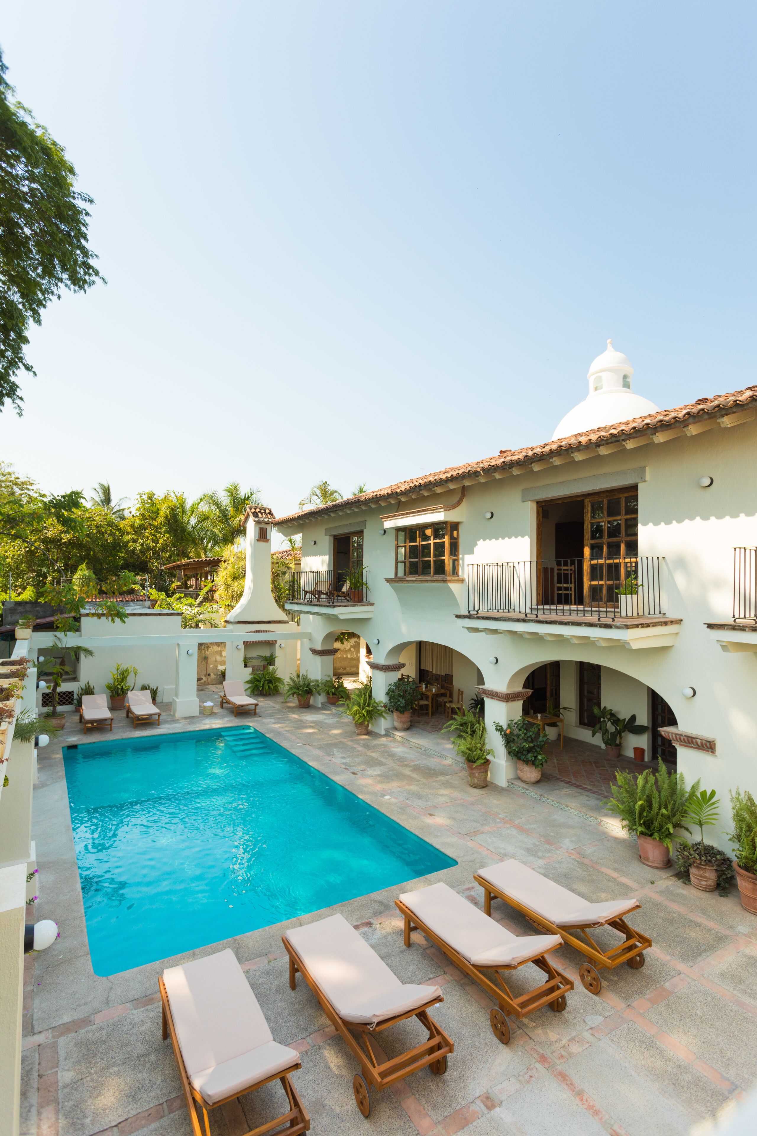 Un área de piscina con sillones de descanso, un edificio blanco con tejado de azulejos y plantas en macetas.