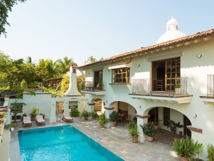 Un área de piscina con sillones de descanso, un edificio blanco con tejado de azulejos y plantas en macetas.