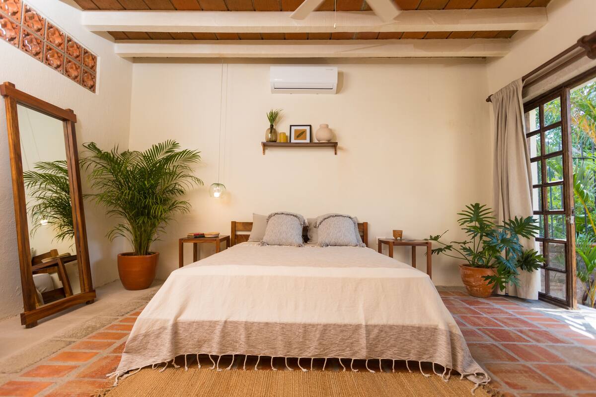 A bedroom with a large bed, a wooden headboard, a potted plant, a mirror, and a window with curtains.