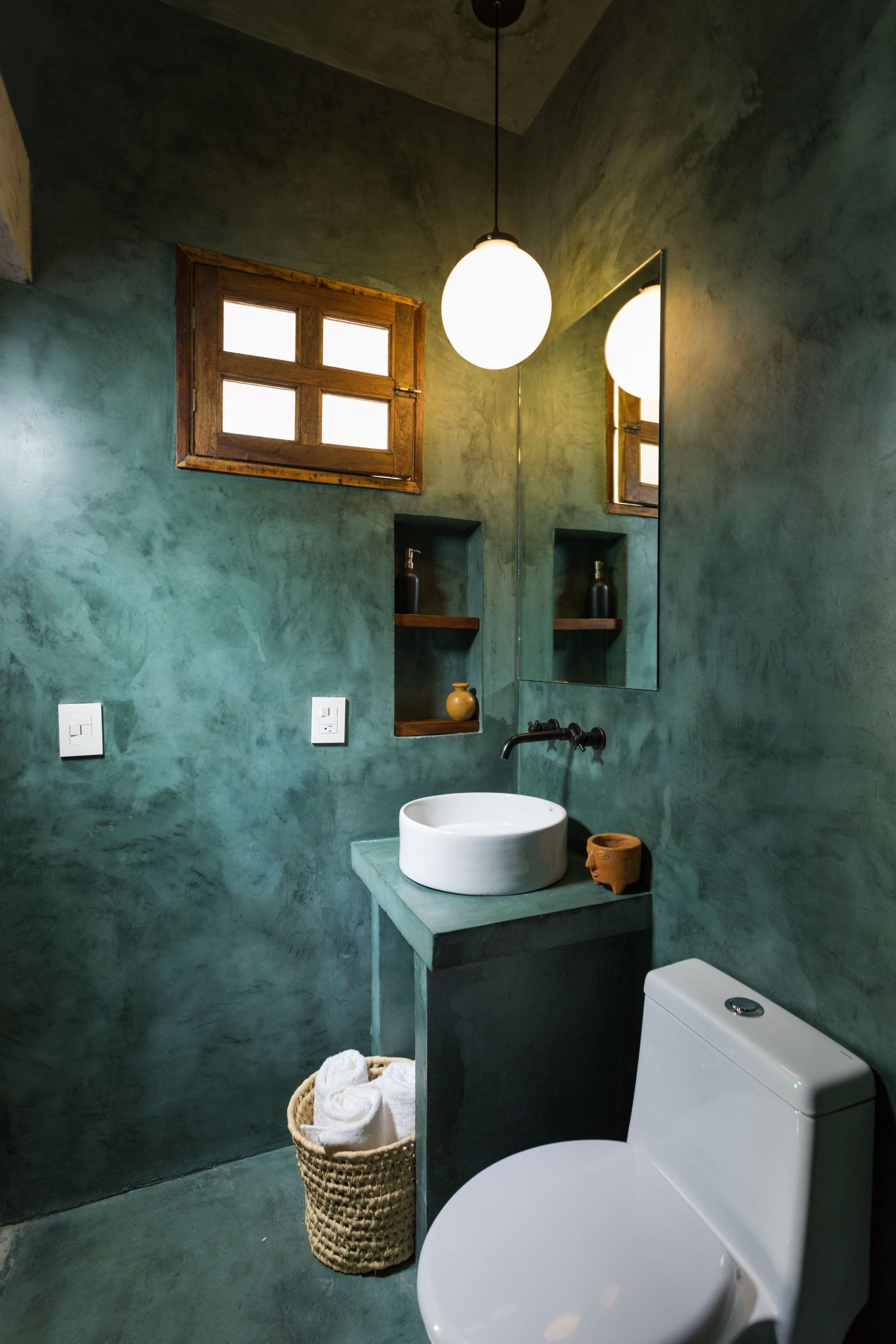 Ein Badezimmer mit rundem Waschbecken, Toilette, einer Pendelleuchte, einem Spiegel und einem Fenster mit Holzjalousien.