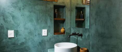 Ein Badezimmer mit rundem Waschbecken, Toilette, einer Pendelleuchte, einem Spiegel und einem Fenster mit Holzjalousien.