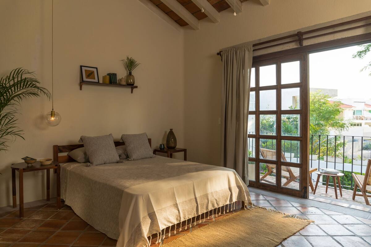 A bedroom with a large bed, a window with a view, a small table, and a potted plant.