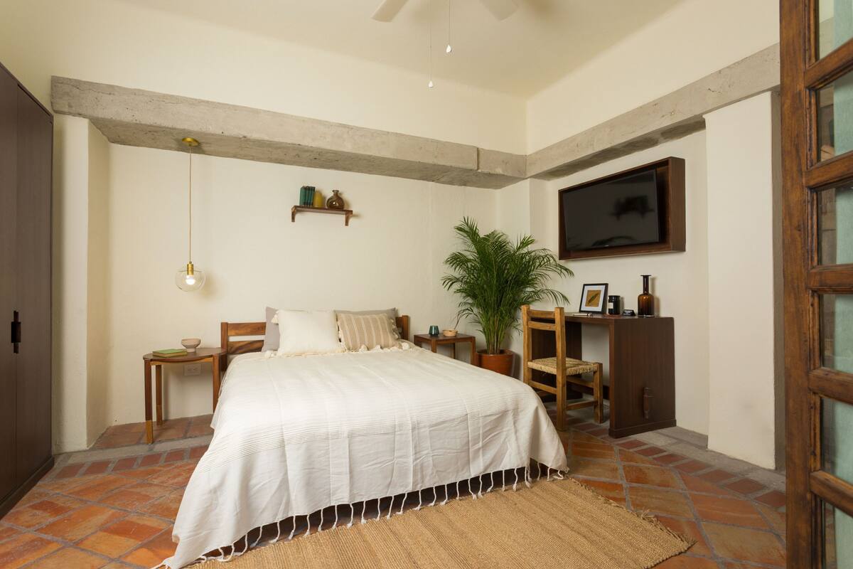 A bedroom with a bed, a desk, a chair, a potted plant, and a television on the wall.