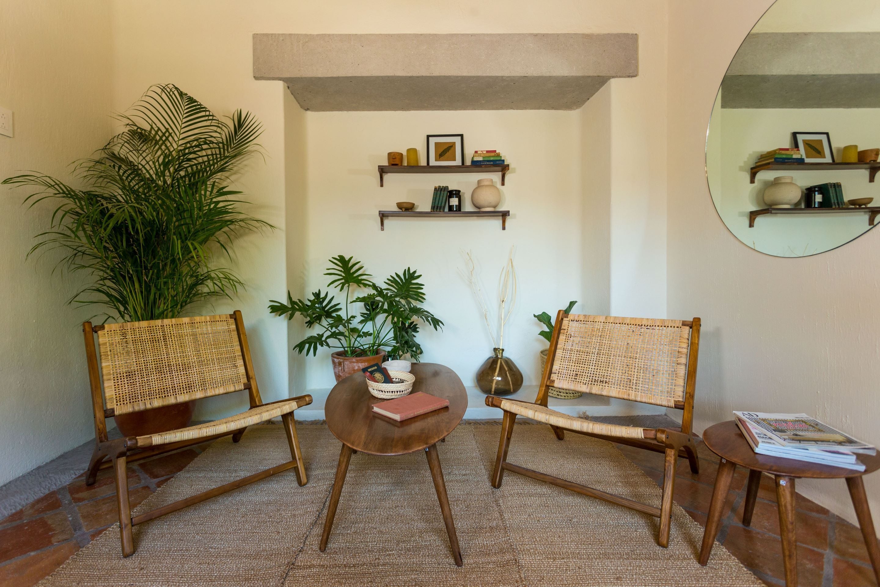Um quarto com duas cadeiras de vime, uma mesa redonda pequena e uma planta em vaso. Há prateleiras com livros e objetos decorativos na parede.