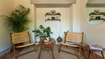 Um quarto com duas cadeiras de vime, uma mesa redonda pequena e uma planta em vaso. Há prateleiras com livros e objetos decorativos na parede.