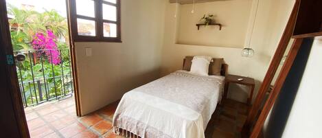 A bedroom with a bed, a small bedside table, a nightstand, a window, and a view of a garden with pink flowers.