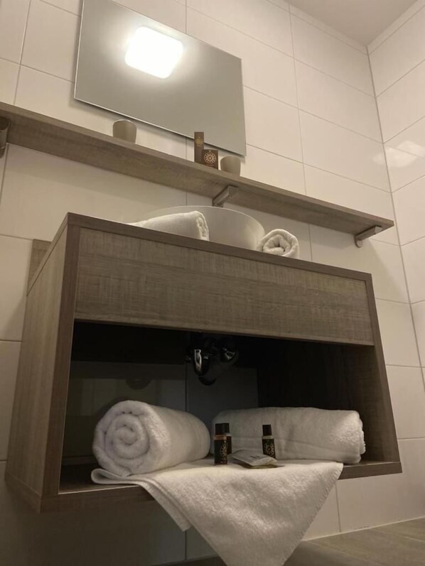 Un baño con tocador de madera, un espejo y estante con toallas y artículos de aseo.
