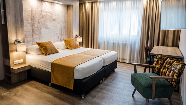Habitación de hotel moderna con una cama grande, una mesita de noche con lámpara, una silla y un amplio ventanal con cortinas.