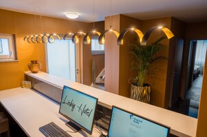 Balcão de recepção de hotel com dois monitores de computador exibindo o nome do hotel e uma planta próxima.