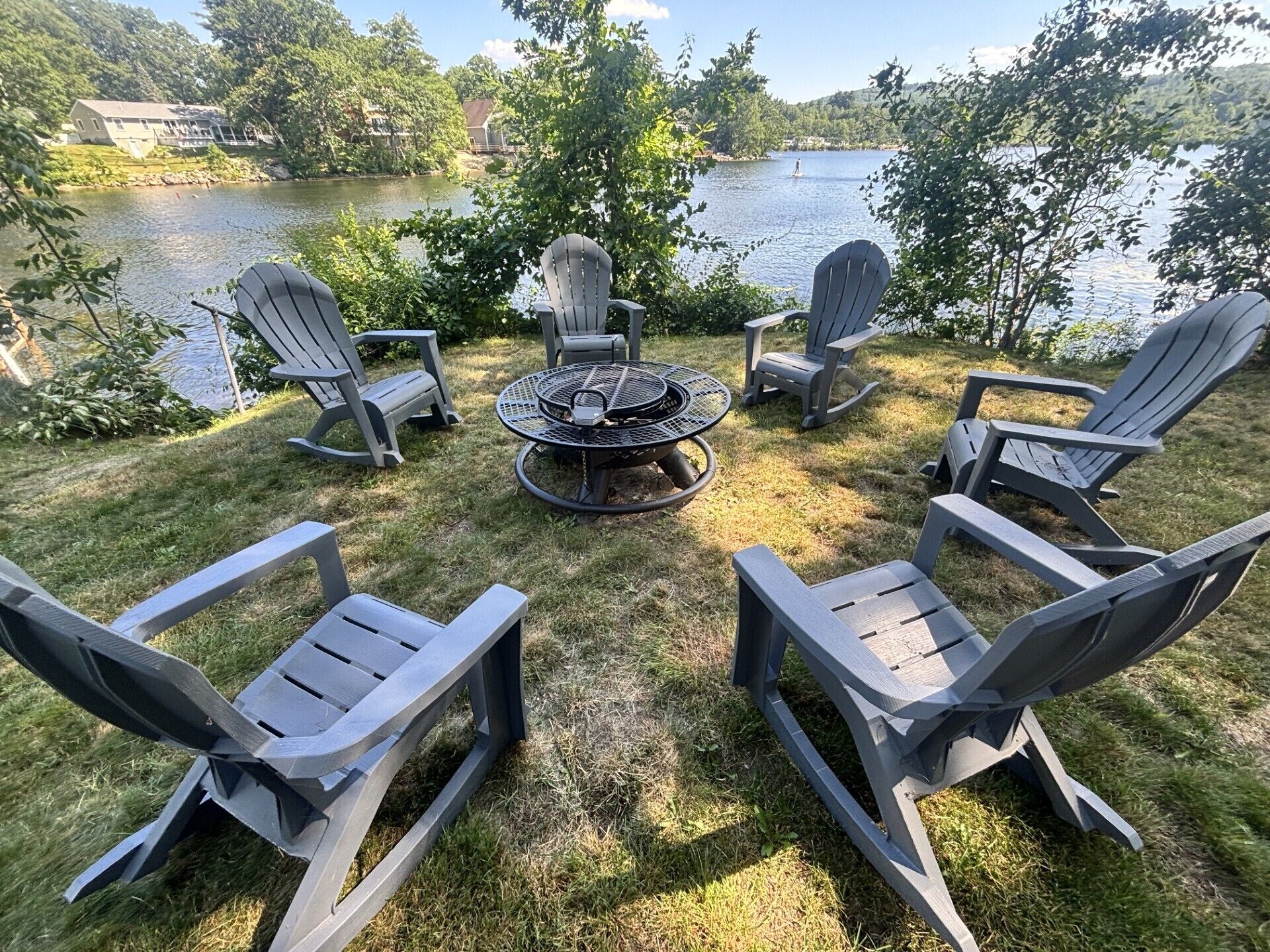 Ein runder Außenbereich mit Adirondack-Stühlen und einem Feuerkorb, umgeben von einer Rasenfläche mit Blick auf einen See.