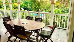 A covered outdoor dining area with a round table and chairs, surrounded by lush greenery and a view of a pool. - Villa Mabouya (6 pers.) (Deshaies)