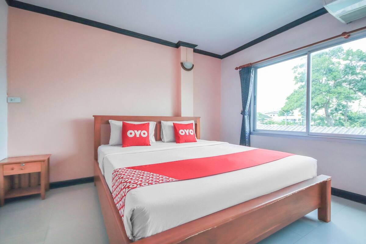 A hotel room with a large bed, two pillows with the brand name "OYO", a red blanket, a wooden nightstand, and a window with a view of trees.