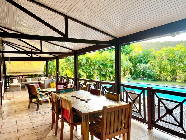 A covered outdoor area with a pool, wooden furniture, and a view of lush greenery.
