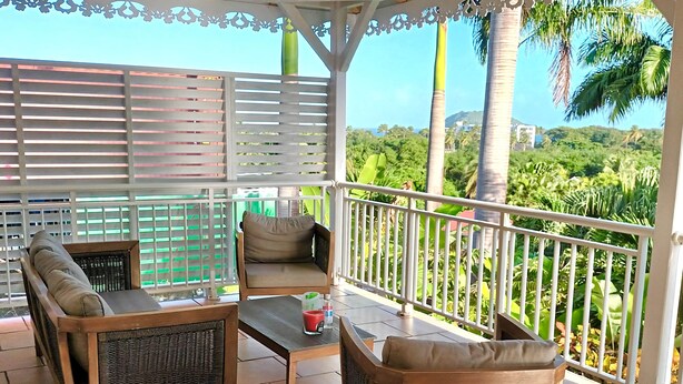 A covered outdoor seating area with wicker furniture, a table, and a view of lush greenery and a distant hill.
