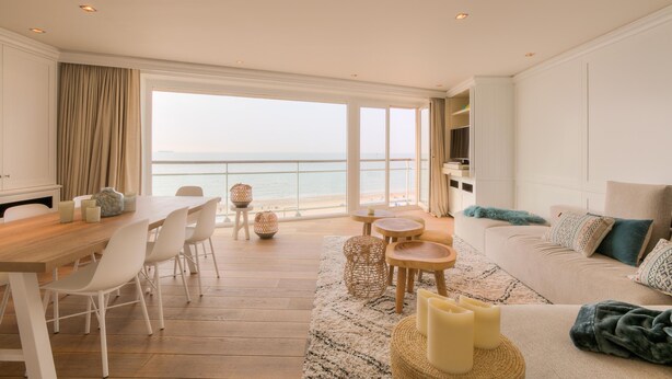Un'ampia zona giorno con un tavolo da pranzo in legno, sedie bianche, un divano color beige e vista sul mare attraverso ampie finestre.