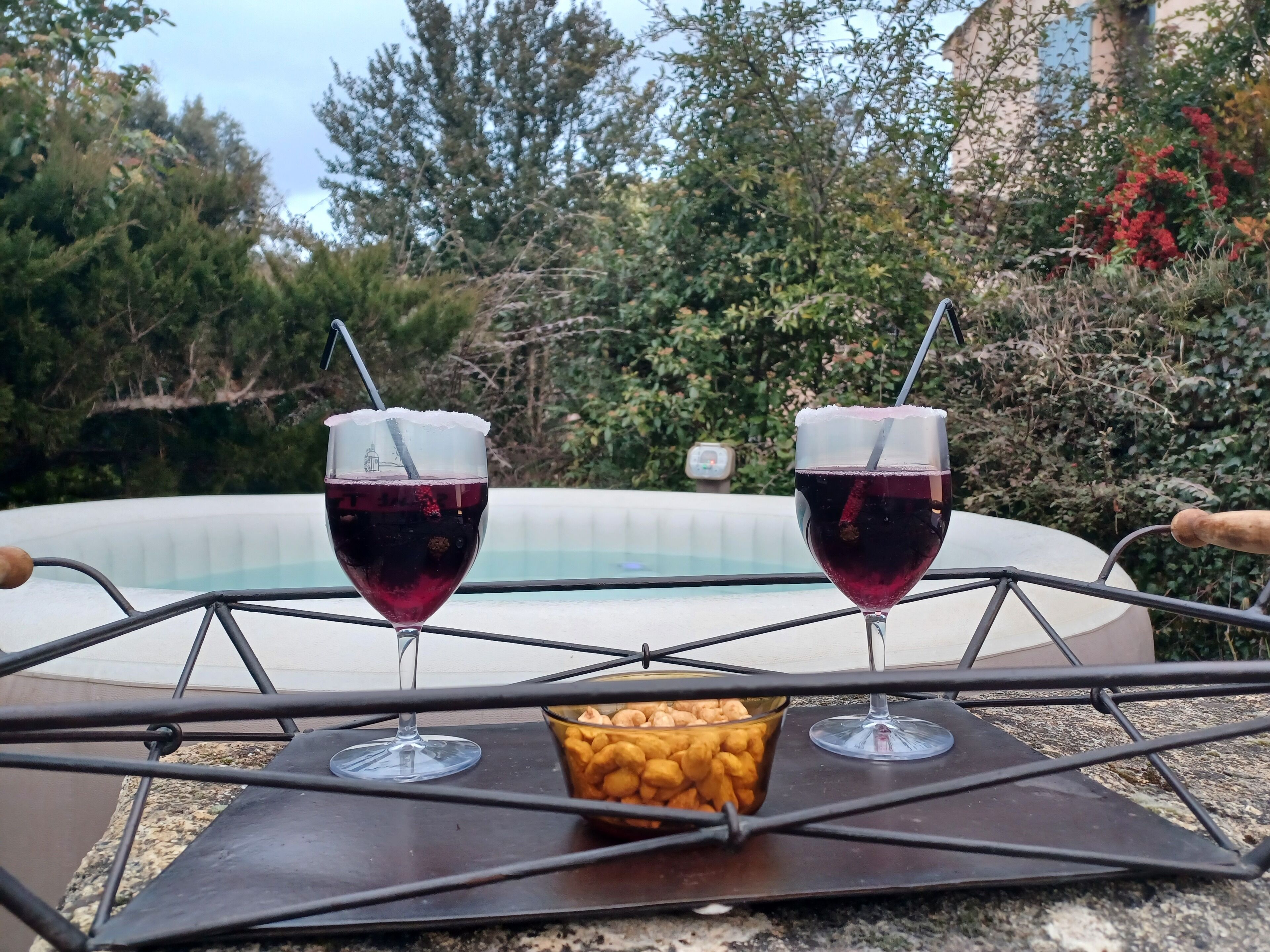 Two glasses of red beverage on a metal tray with a bowl of snacks, in front of a hot tub.
