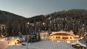 A snow-covered lodge with a central building surrounded by snow-laden trees, a fenced area, and a body of water in the foreground. - Luxury Penthouse Condo (Sun Peaks)