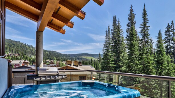 A hot tub on a balcony with a view of a forest and houses. - Luxury Penthouse Condo (Sun Peaks)