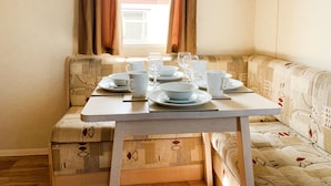 A dining area with a table set for four, a cushioned bench, and a window with curtains. - Beautiful 3-bedroom Caravan at Mersea Island (Colchester)