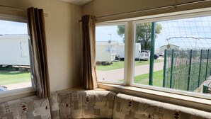 A corner seating area with a patterned fabric, a window with curtains, and a view of a fenced outdoor area with vehicles. - Beautiful 3-bedroom Caravan at Mersea Island (Colchester)