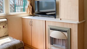 A compact room with a television on a shelf, a patterned sofa, and a built-in heater. - Beautiful 3-bedroom Caravan at Mersea Island (Colchester)