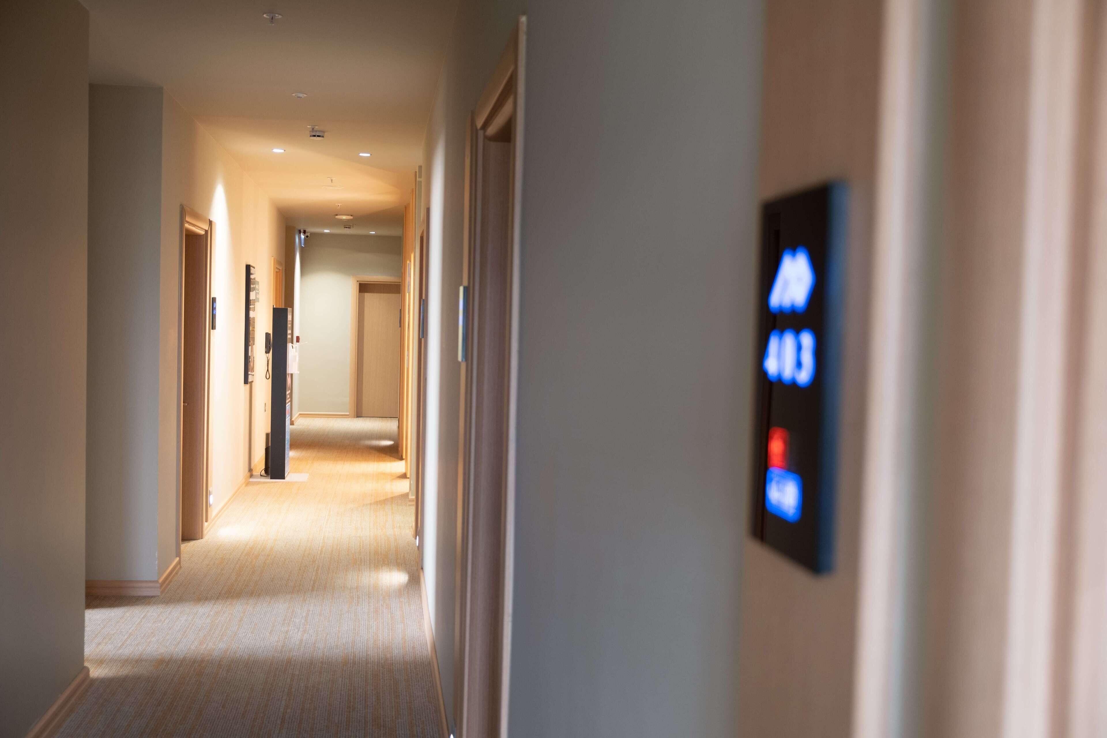 A hotel hallway with a carpeted floor, a door on the left, and a digital display on the right.