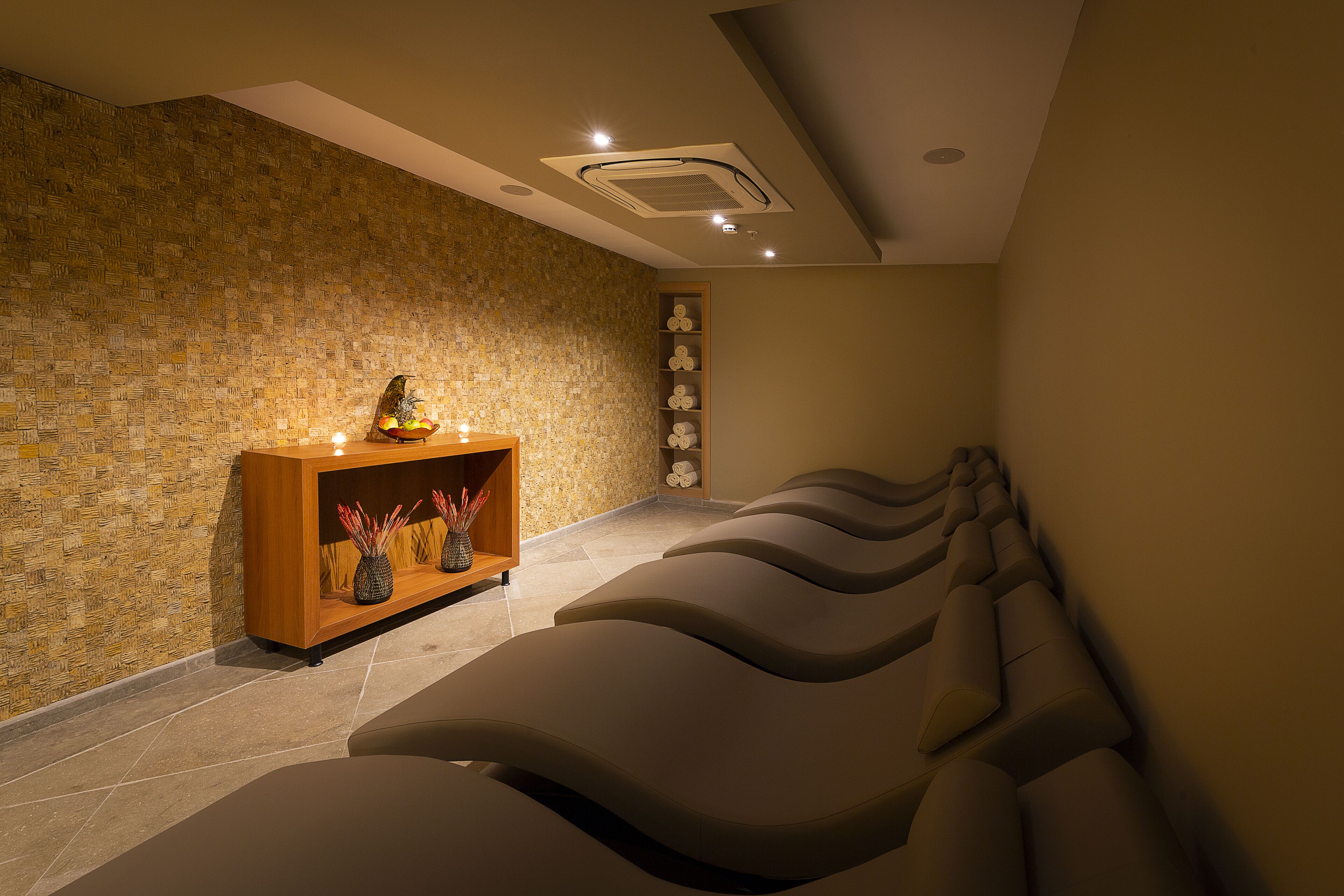 A spa room with a stone wall, a wooden shelf with decorative items, and a row of massage chairs.