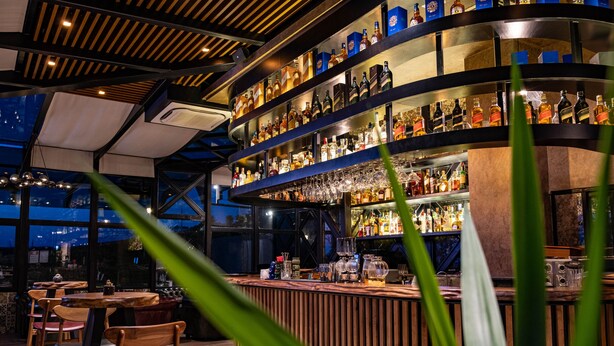 Zona de bar con estantes llenos de botellas de licor y vasos, sillas de madera y un diseño moderno en el techo.