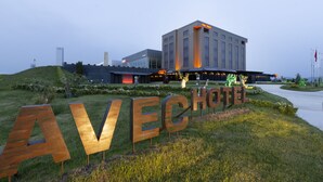 Un letrero de hotel con el nombre "AVECHOTE" en letras grandes, un edificio moderno con fachada de vidrio y un área ajardinada con césped y flores.