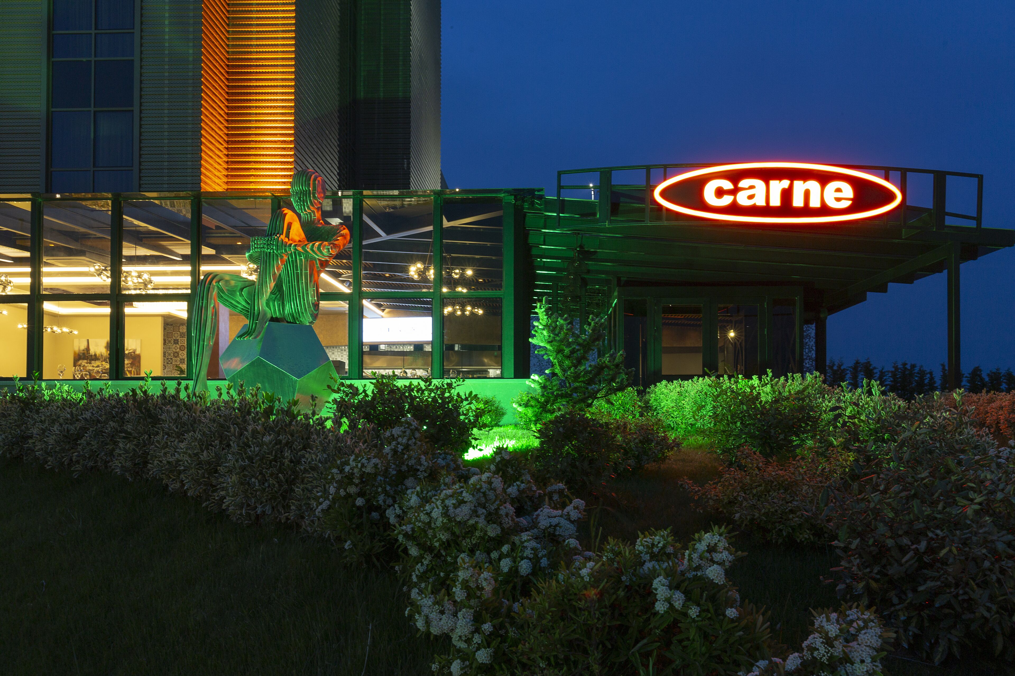 A building with a 'carne' sign, large glass windows, and a green-lit sculpture in front.
