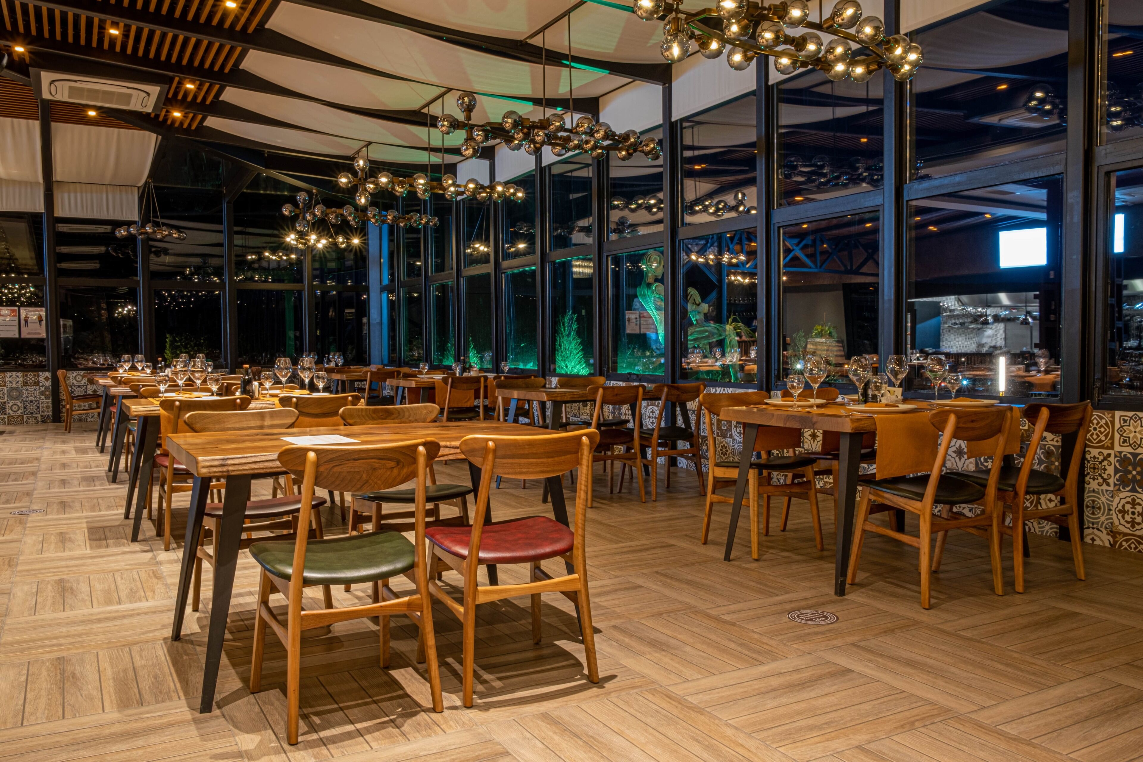 A restaurant with wooden tables and chairs, large windows, and hanging light fixtures.