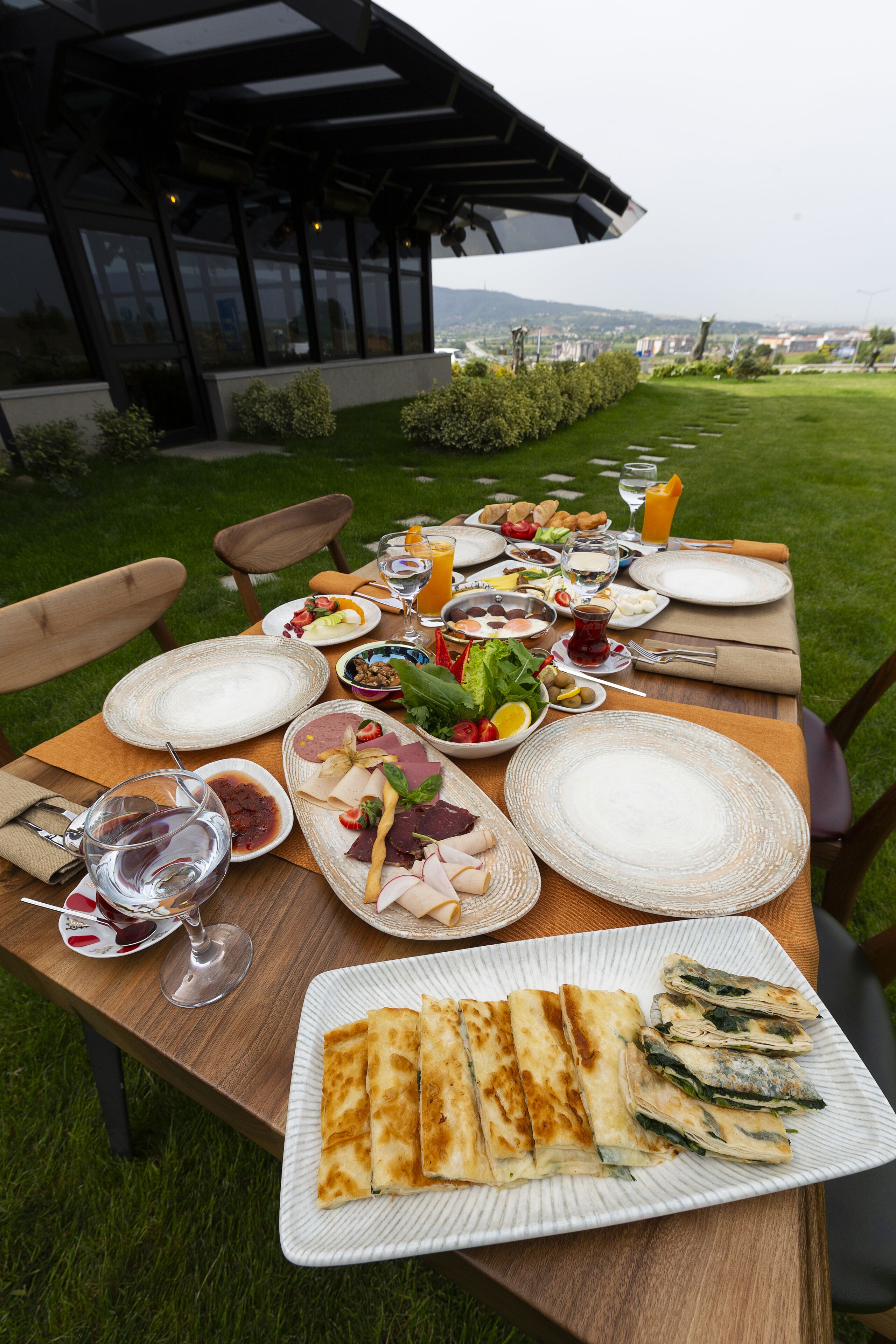 Mesa exterior dispuesta con diversos alimentos, copas de vino y una vista panorámica de fondo.