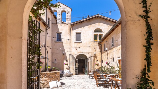 An arched entrance leading to a cobblestone courtyard with stone walls, a wooden table, and potted plants. - Relais Convento Santa Croce (Sant'Anatolia di Narco)